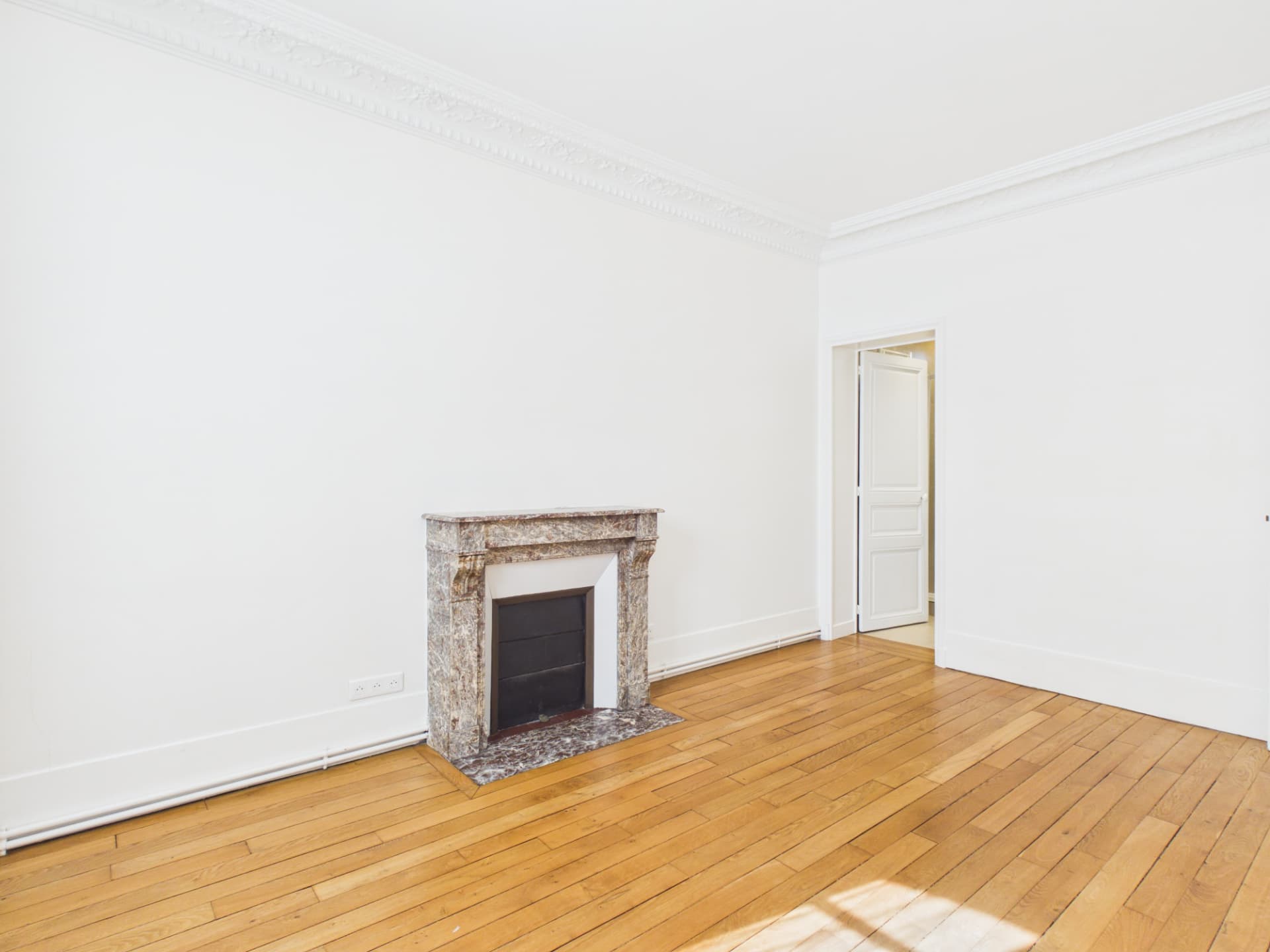Pièce parisienne vide et lumineuse avec parquet, moulures et cheminée en marbre